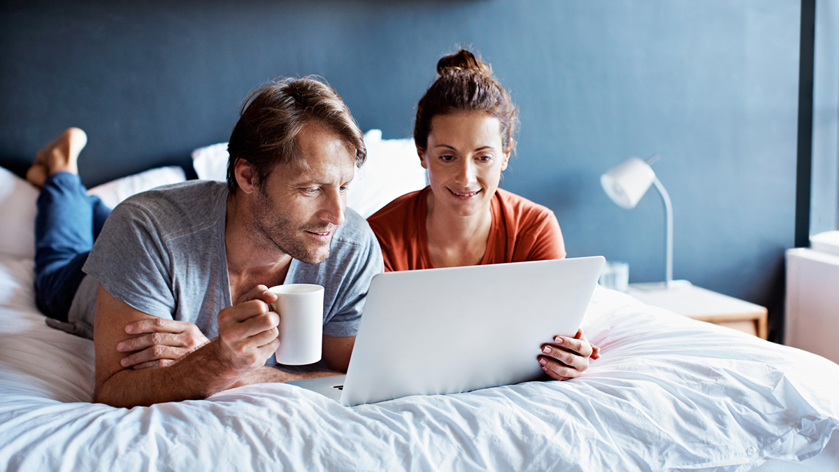 Par som ligger på sengen med kaffe og nettbrett. Foto.
