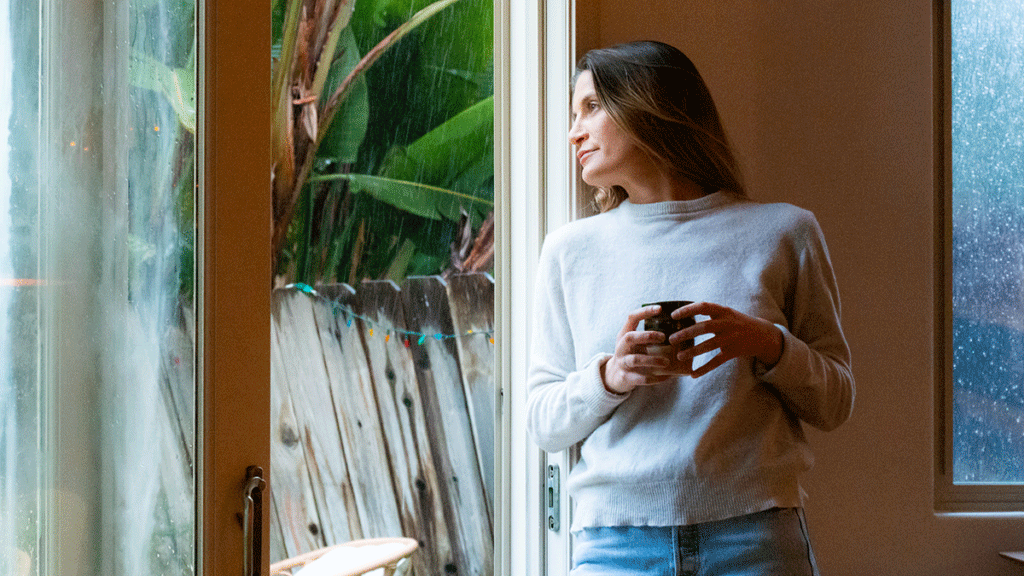 Dame med kaffekopp står inne og titter ut på regnet. Foto.